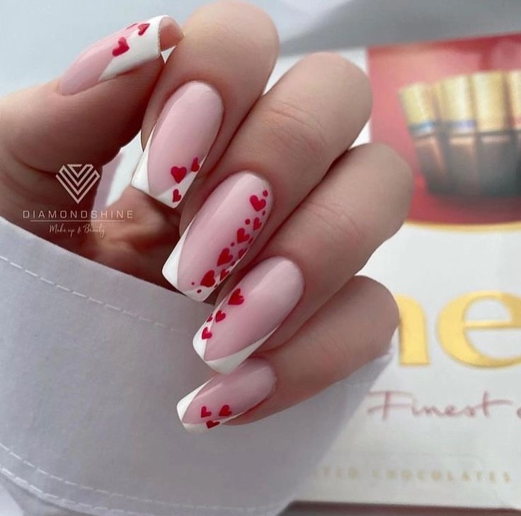 Hands-with-Cupids-coffin-nails-featuring-slanted-white-French-tips-and-cascading-red-hearts-on-a-blush-pink-base-paired-with-a-white-shirt.jpg